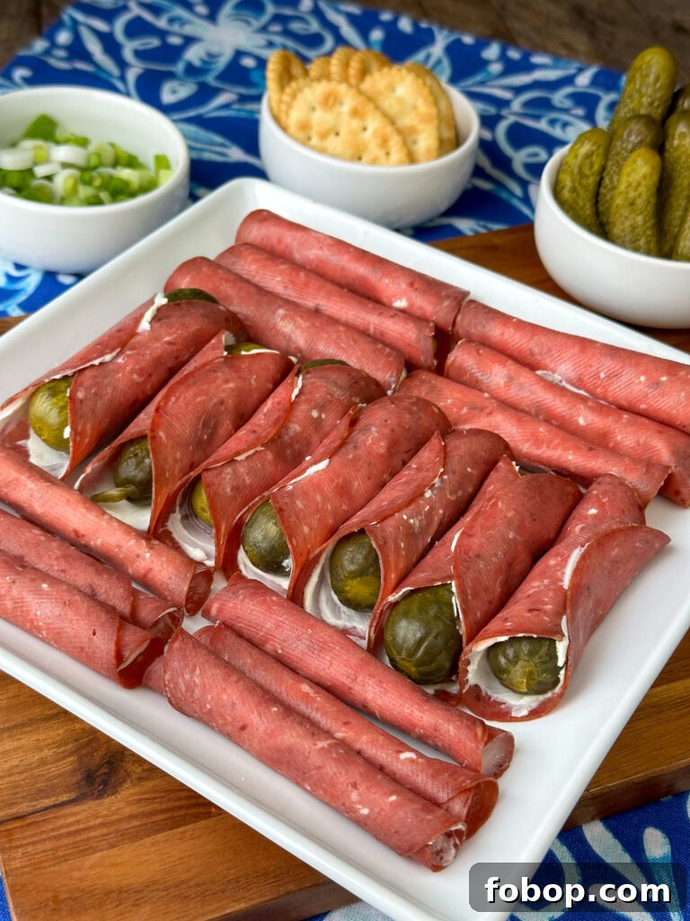 Savory Dried Beef Roll-Ups Recipe for Appetizers and Snacks 3 A row of dill pickles wrapped in a cream cheese and beef roll-up surrounded by perpendicular rows of plain dried beef and cream cheese roll-ups on a square white plate with crackers and pickles behind on a blue and white napkin.
