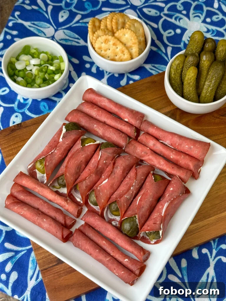 Savory Dried Beef Roll-Ups Recipe for Appetizers and Snacks 8 Overhead view of a white platter with rows of dried beef and cream cheese roll-ups in a decorative pattern sitting on a brown cutting board with small dishes of garnishes beside the board.