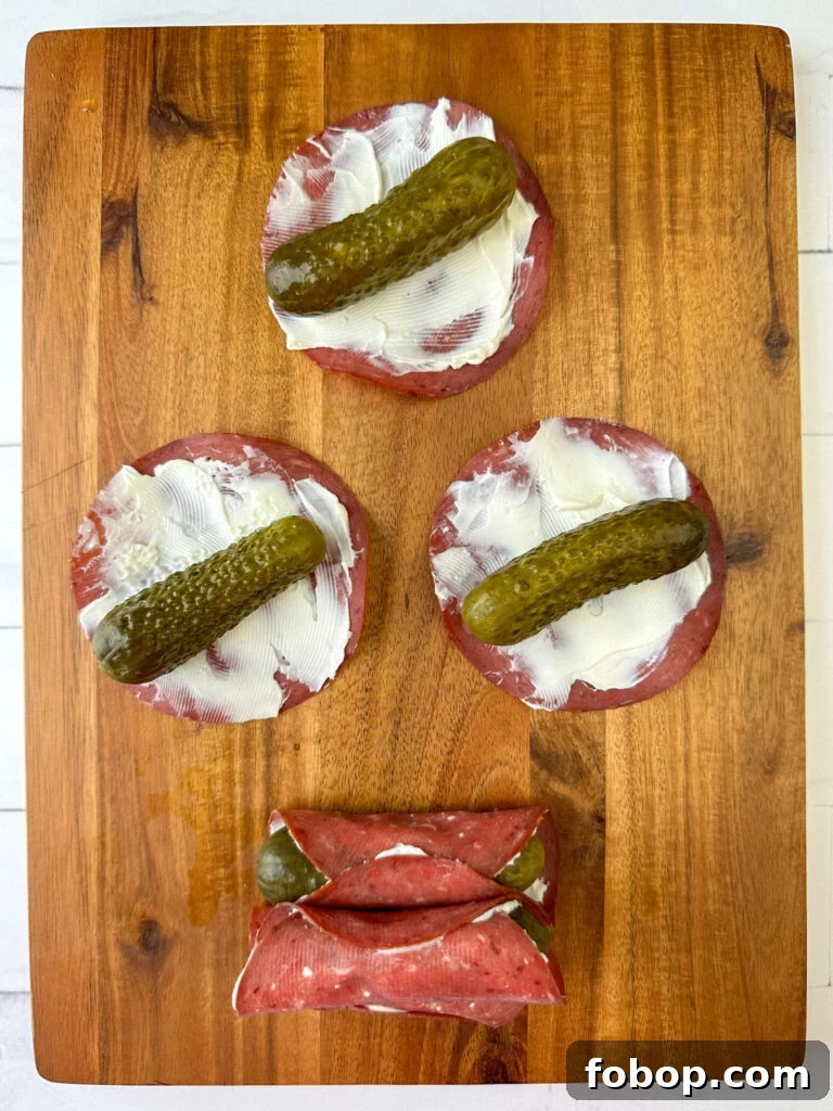 Savory Dried Beef Roll-Ups Recipe for Appetizers and Snacks 9 Baby dill pickle laying on a thin layer of cream cheese lined dried beef rounds on a brown cutting board.