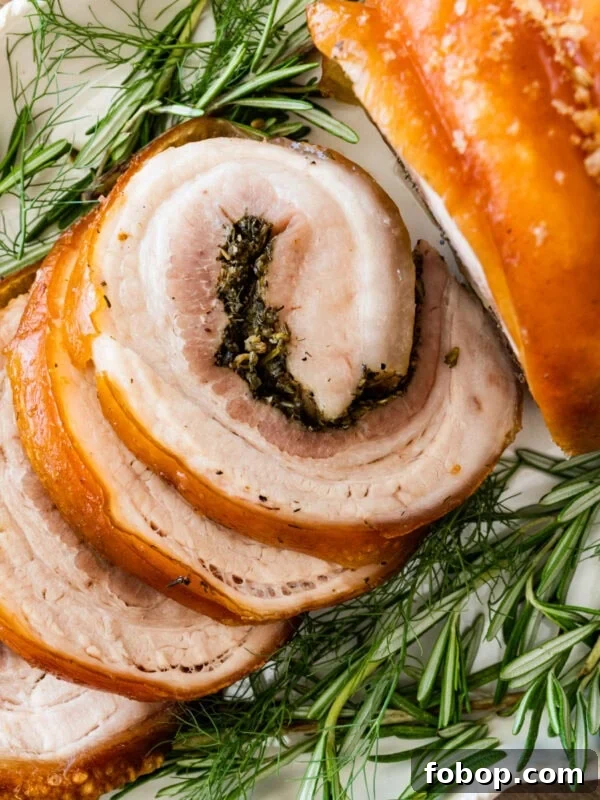 Beautiful porchetta roast with crispy skin and juicy interior and herb layer on a platter ready to serve. Fresh herbs around the white platter.