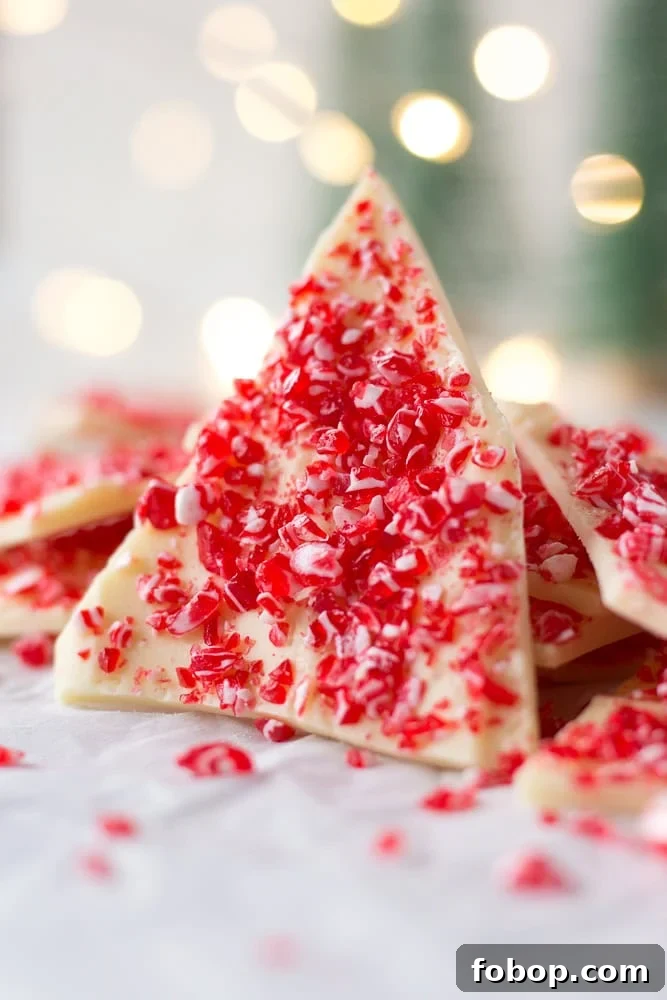Gluten-Free White Chocolate Peppermint Bark Recipe 2 A triangular piece of white chocolate peppermint bark standing up against other pieces of peppermint bark. Twinkly lights are in the background.