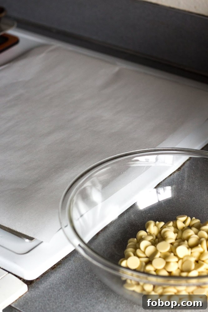 Gluten-Free White Chocolate Peppermint Bark Recipe 3 A piece of white parchment on a sheet pan next to a clear bowl with white chocolate chips.