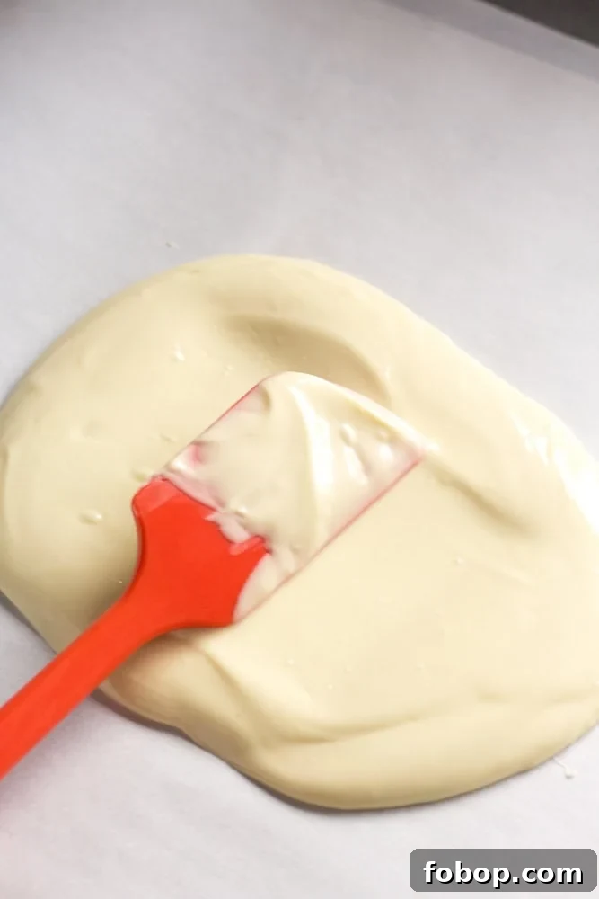 Gluten-Free White Chocolate Peppermint Bark Recipe 8 A red spatula spreading white chocolate on a sheet pan lined with parchment paper.