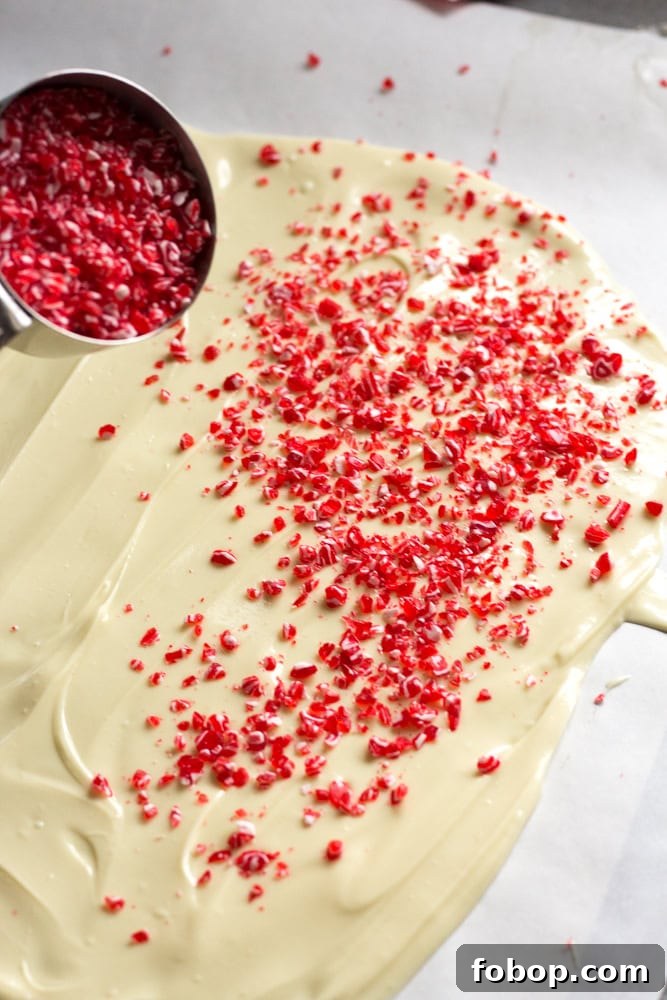 Gluten-Free White Chocolate Peppermint Bark Recipe 10 Red and white bits of peppermint candy being poured out of a gray measuring cup onto a slab of melted white chocolate.