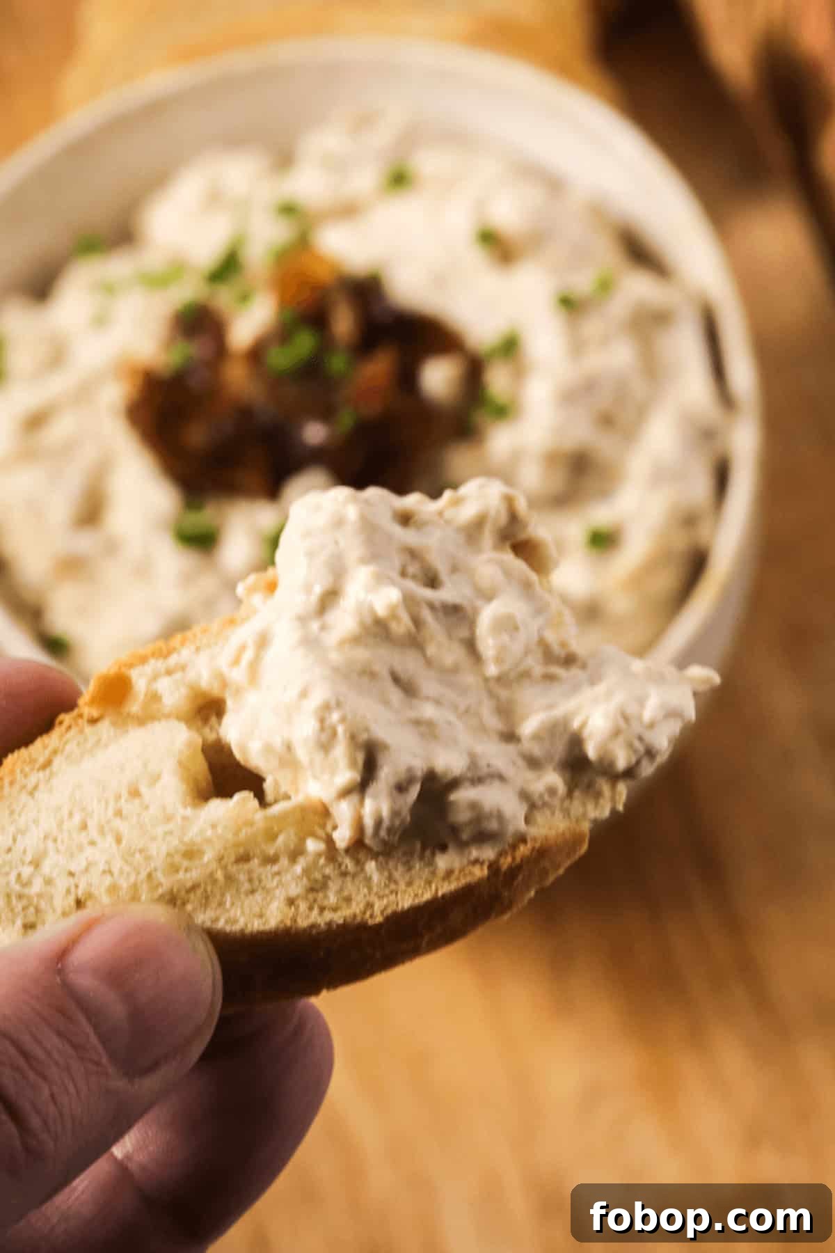 A hand holds a piece of bread topped with creamy caramelized onion dip. In the background, a bowl of dip garnished with chopped chives and sweet caramelized onions is visible.