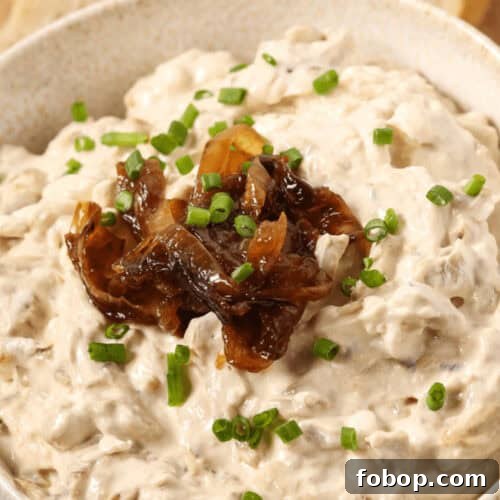 A bowl of rich caramelized onion dip is topped with chopped chives, with sliced bread in the background.