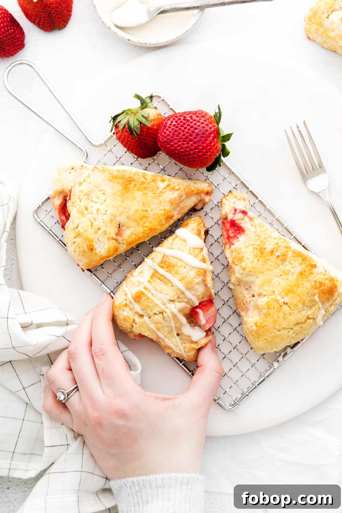 Woman's hand reaching to take a homemade strawberry scone. Scone is on wire holder with 2 other scones and 2 fresh strawberries.