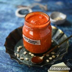 Smoky Roasted Red Pepper Pesto Recipe for Pasta & Sandwiches 8 Red pepper pesto in a jar with grated cheese in the front on a blue surface.