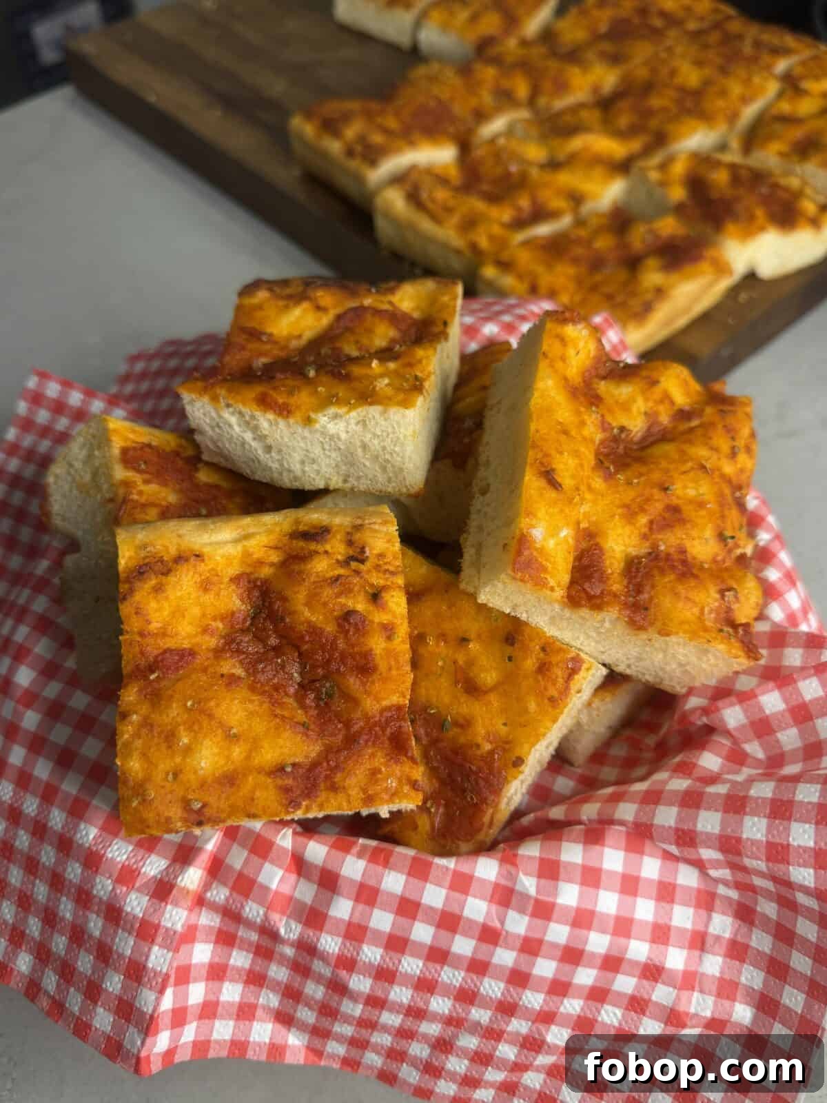 Carbone Fine Foods Grandma-Style Italian Bread 4 A basket lined with red and white checkered cloth holds several square slices of focaccia bread topped with tomato sauce, with more slices on a wooden board in the background.