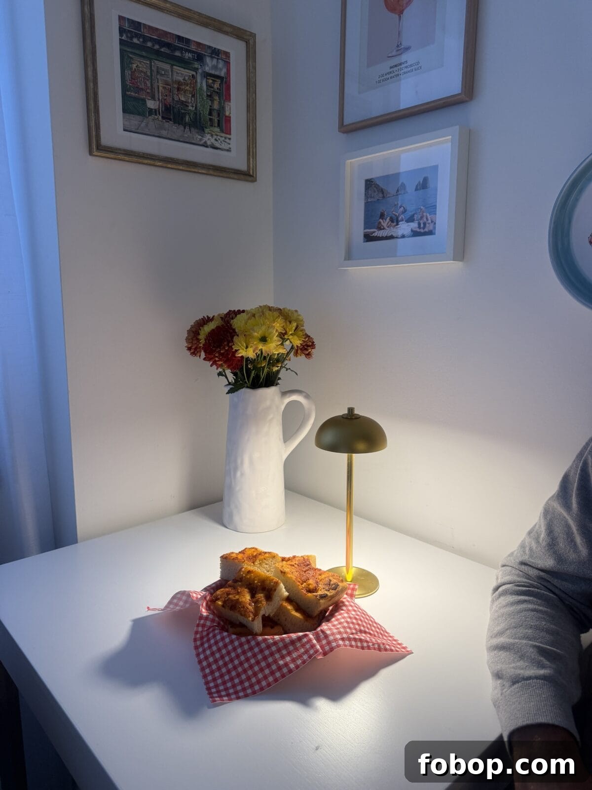 Carbone Fine Foods Grandma-Style Italian Bread 5 A small white table with a plate of pastries on a red checkered napkin, next to a gold lamp and a white pitcher with red and yellow flowers. Framed art hangs on the wall above.
