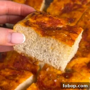 Carbone Fine Foods Grandma-Style Italian Bread 6 A hand with manicured nails holds a thick, square slice of focaccia bread topped with tomato sauce and herbs. Other pieces of the same focaccia are visible in the background.