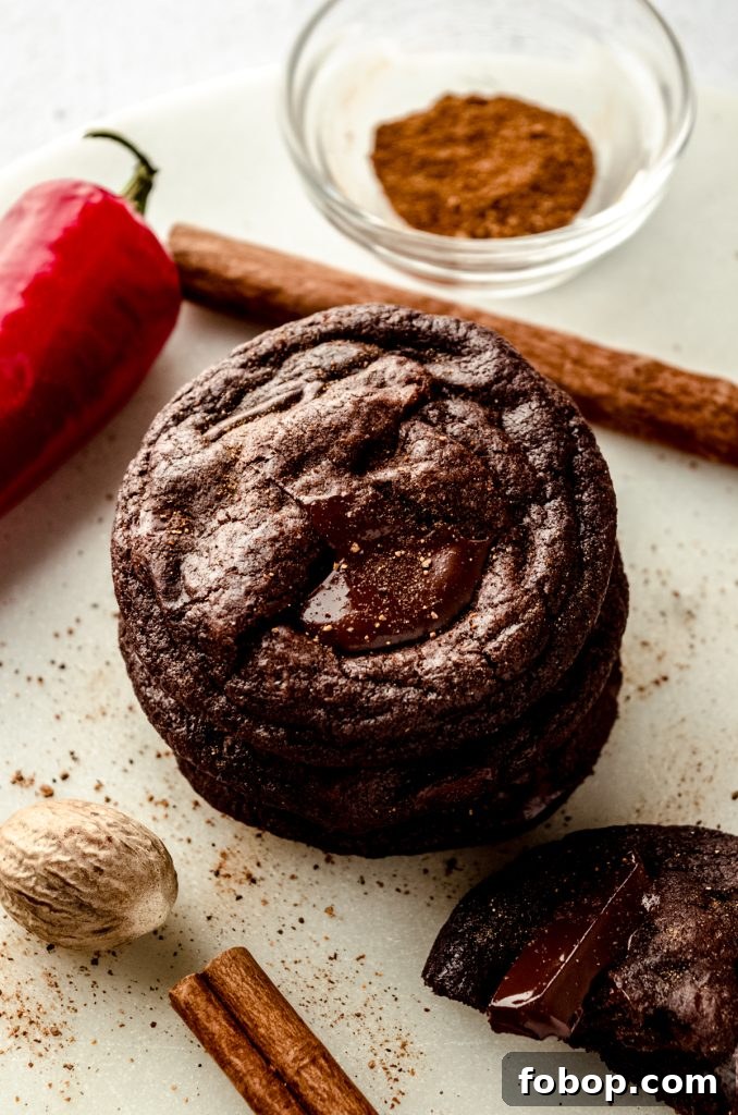 A stack of chocolate cayenne cookies with chili peppers, nutmeg, and cinnamon sticks around it.