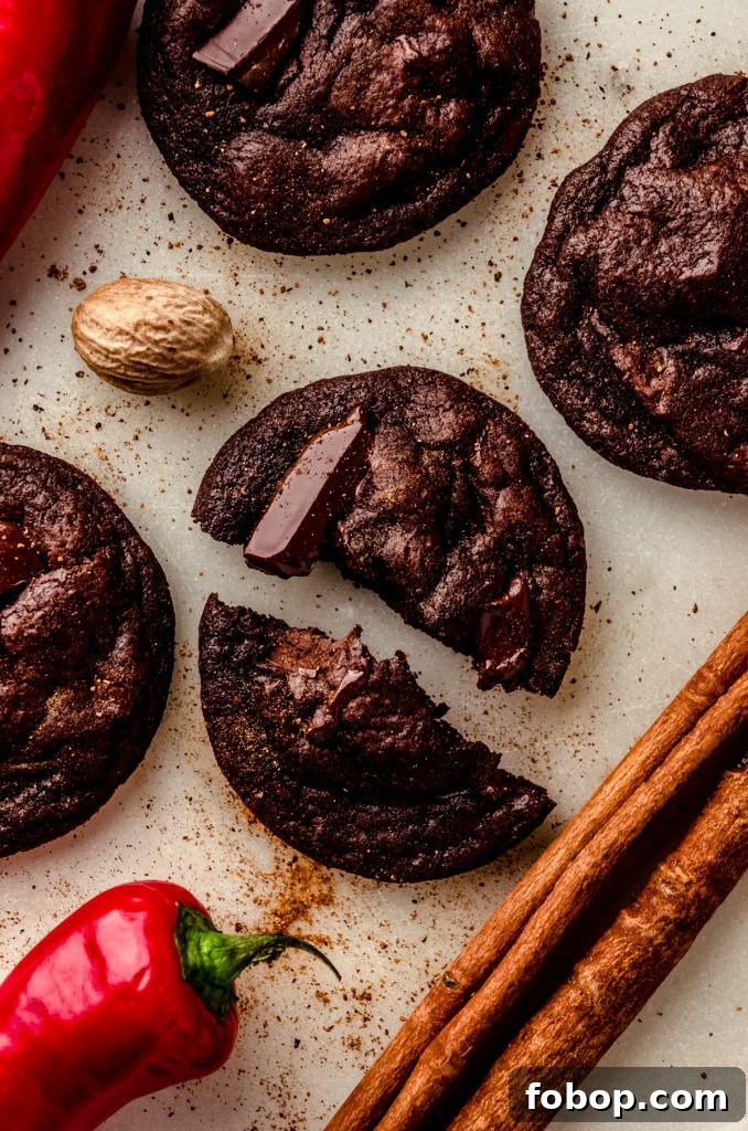 Aerial photo of a chocolate cayenne cookie that has been split in half with a cinnamon stick, chili pepper, and nutmeg around it.