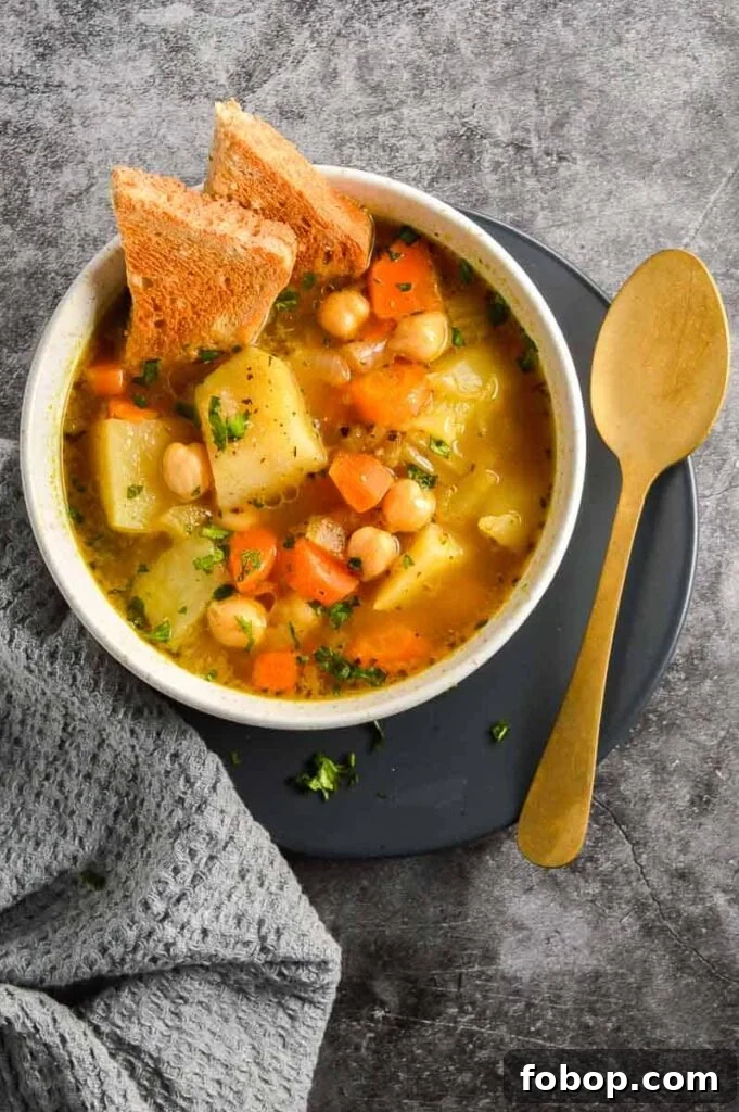Comforting Vegan Potato and Chickpea Stew Recipe 3 Top down view of a pale ceramic bowl with stew and toast in it beside a gold spoon on a grey background