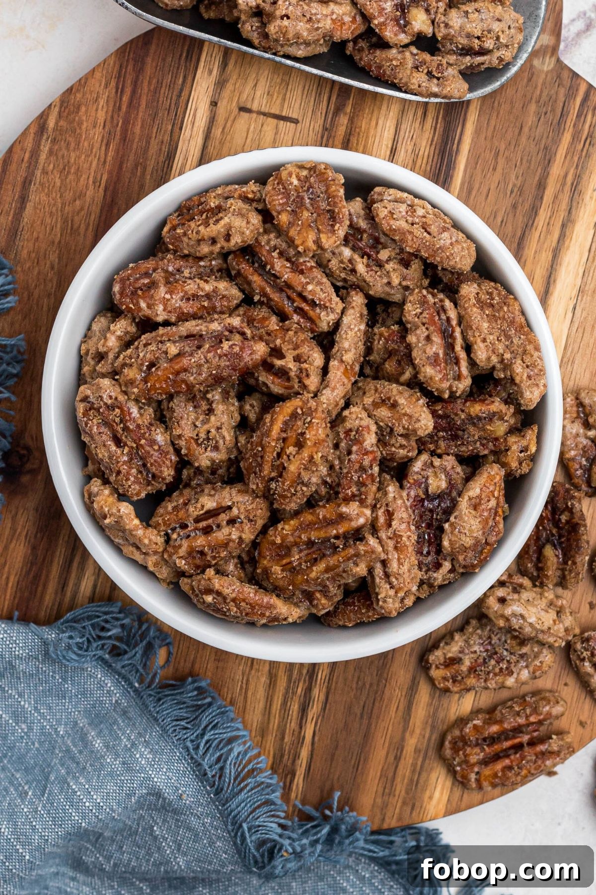 Candied pecans in a small white bowl, with a blue linen and scattered pecans nearby. 