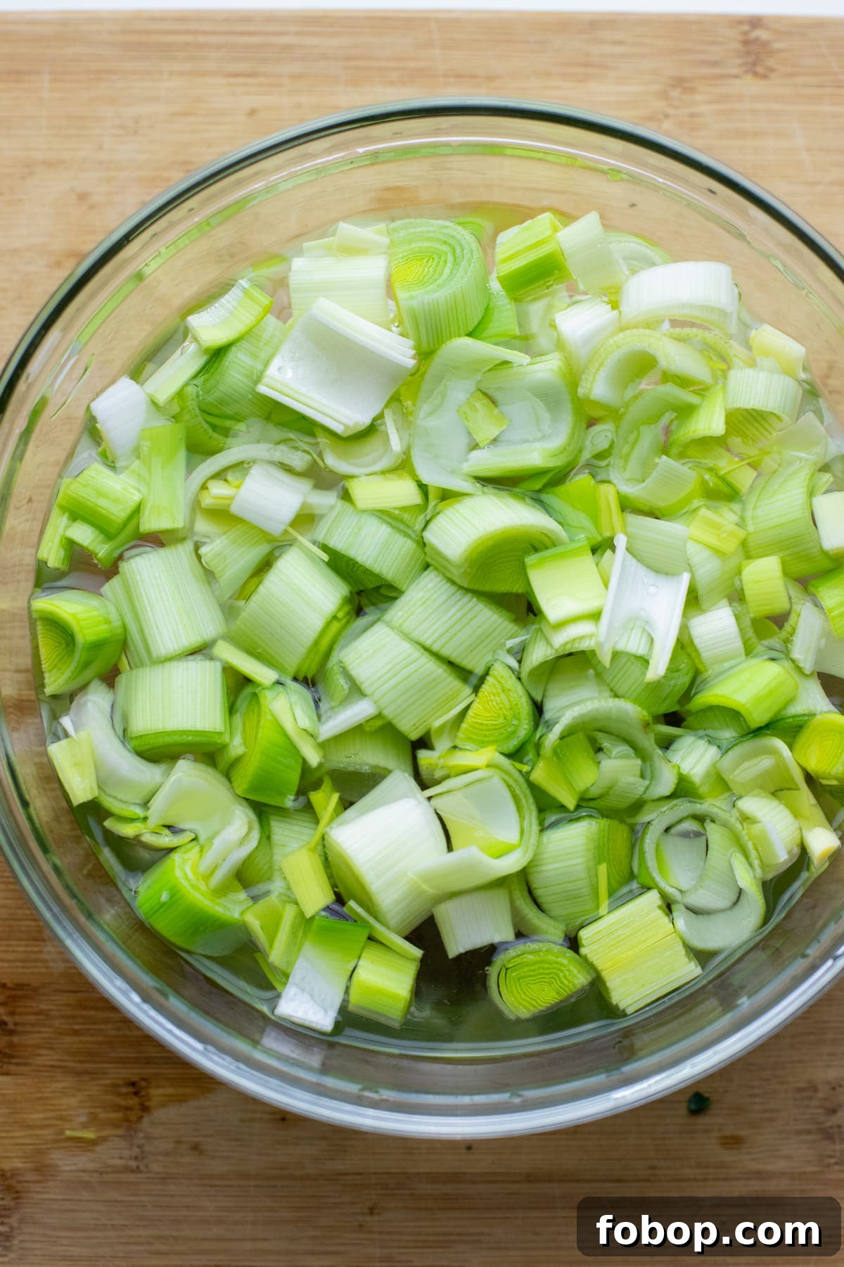 Creamy Vegan Cauliflower Soup Recipe — Dairy-Free Comfort Bowl 3 A clear glass bowl with chopped leeks soaking in water with a cutting board in the background.