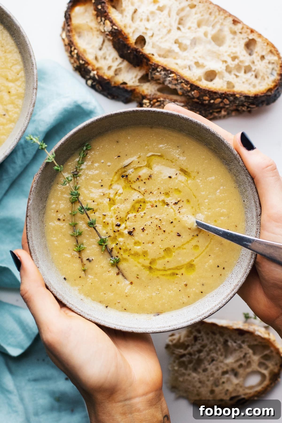 Creamy Vegan Cauliflower Soup Recipe — Dairy-Free Comfort Bowl 8 Two hands holding a grey bowl of cauliflower soup with a blue napkin and bread slices in the background.