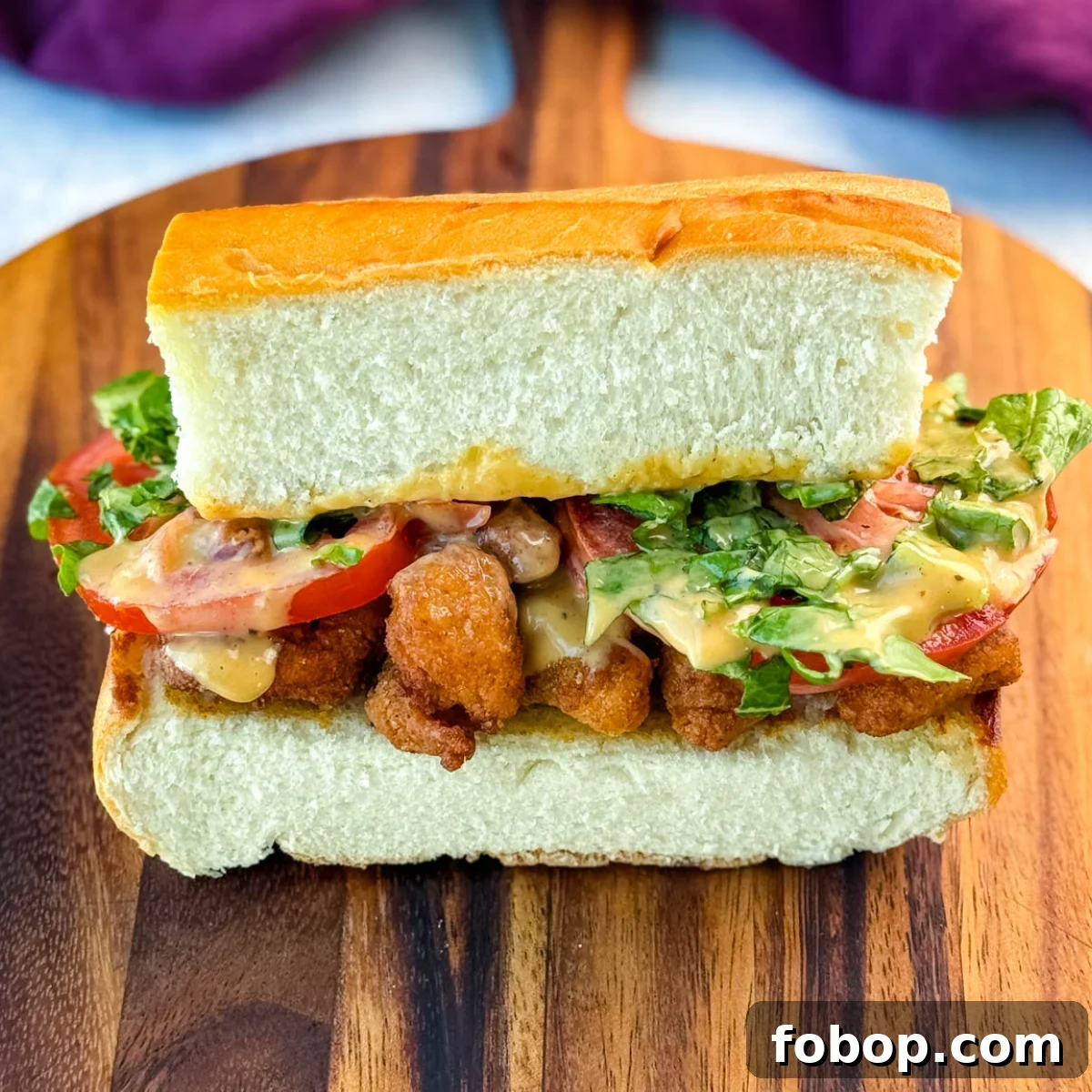 Centered image of a crispy chicken po' boy sandwich on a cutting board, showcasing layers of fried chicken, tomato, lettuce, and sauce, surrounded by chips.
