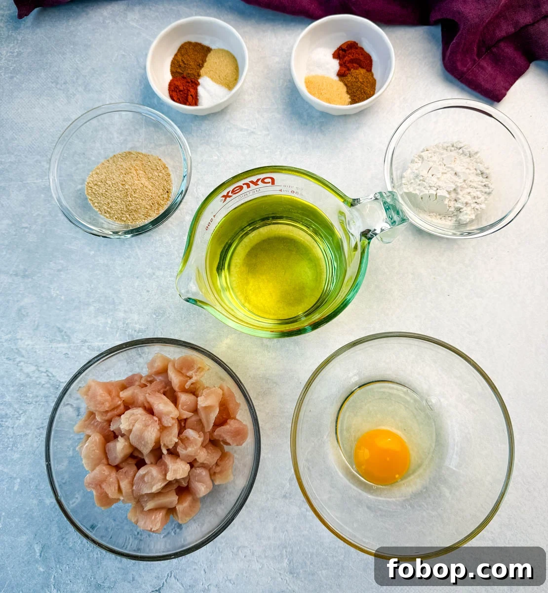 Overhead view of individual ingredients for fried chicken: chopped raw chicken, an egg, flour, breadcrumbs, seasonings, and oil.