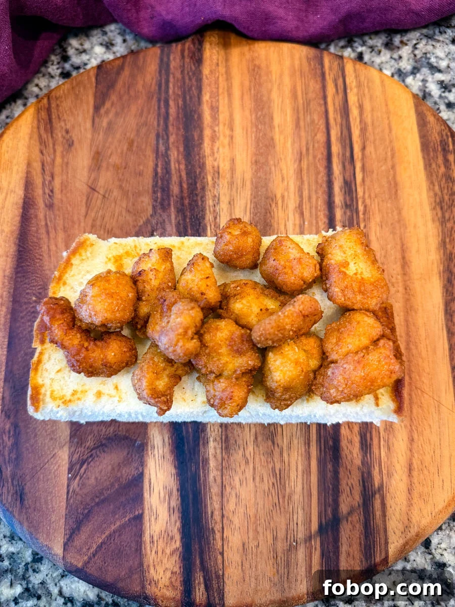 Top-down image showing the bottom half of a sandwich roll loaded with golden, crispy chicken pieces on a wooden board with a granite countertop background.