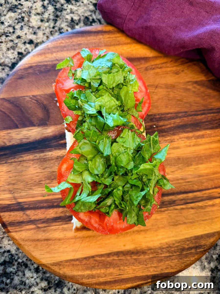 Overhead shot of a sandwich roll topped with fresh tomato slices and shredded romaine lettuce, on a wooden board with a purple napkin nearby.