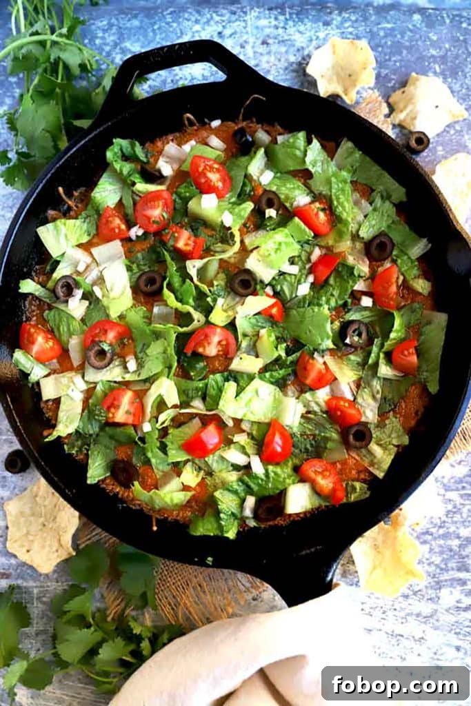 View of a skillet with taco dip topped with lettuce, tomatoes and olives.