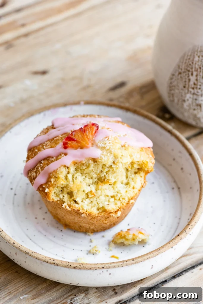 Blood Orange Coconut Muffins with Zesty Glaze 2 A single orange coconut muffin topped with an orange vanilla glaze and a small orange slice on a plate, with a bite missing.