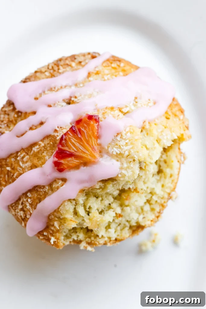 Blood Orange Coconut Muffins with Zesty Glaze 3 Overhead view of an orange muffin with pink glaze and an orange slice on top of a white plate.