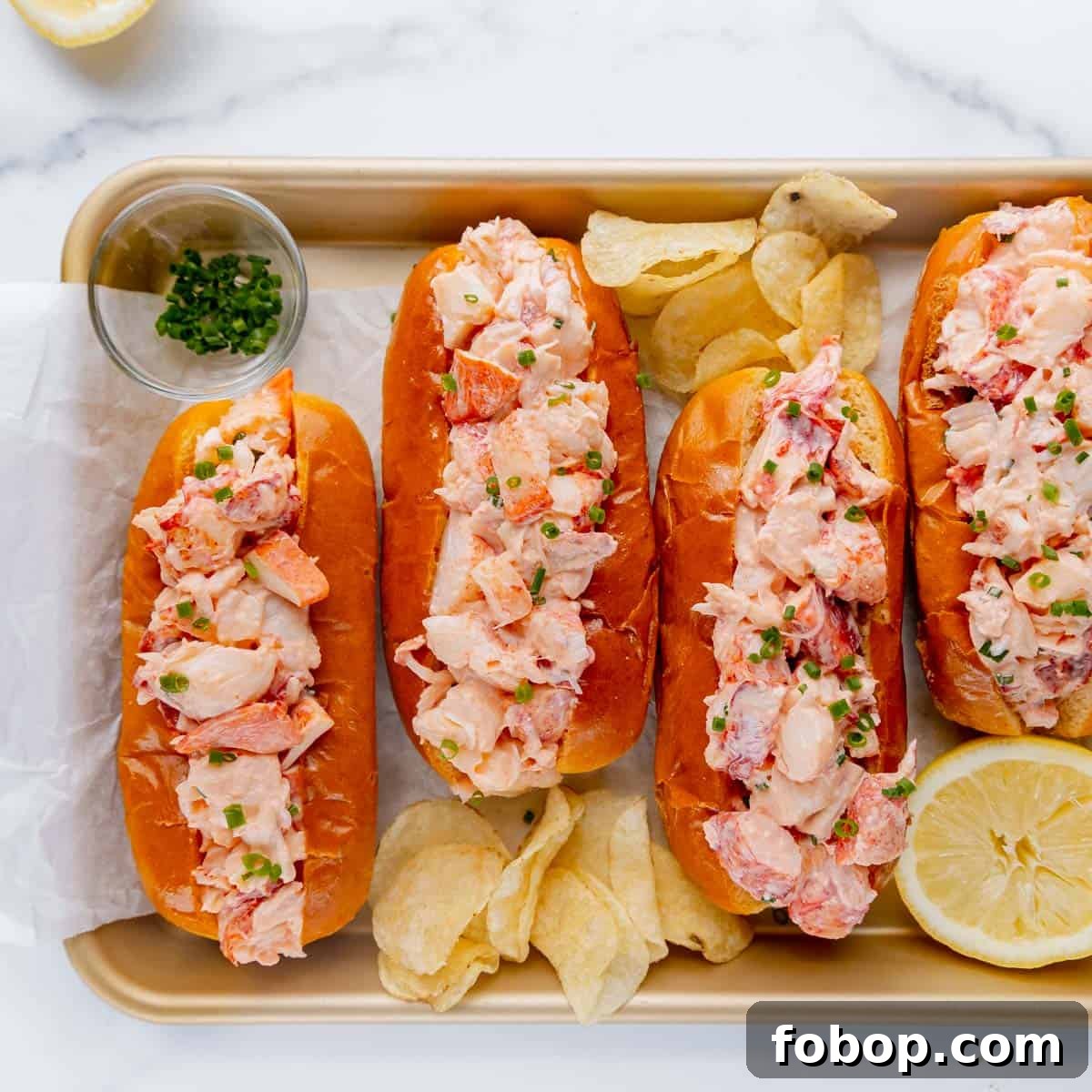 Classic Maine Lobster Roll Recipe: How to Make Authentic Lobster Rolls 3 Four lobster rolls on a tray with chips, lemon and chives.