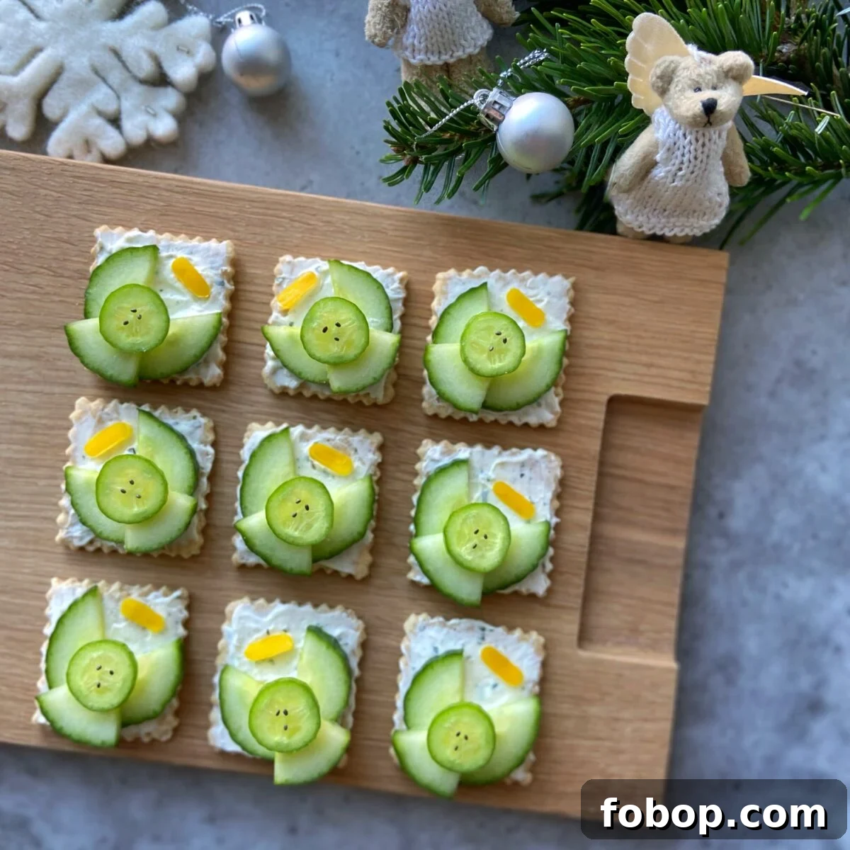 Nine square crackers topped with cream cheese, cucumber slices, and small yellow pepper halos. They are arranged on a wooden board with holiday decorations nearby.
