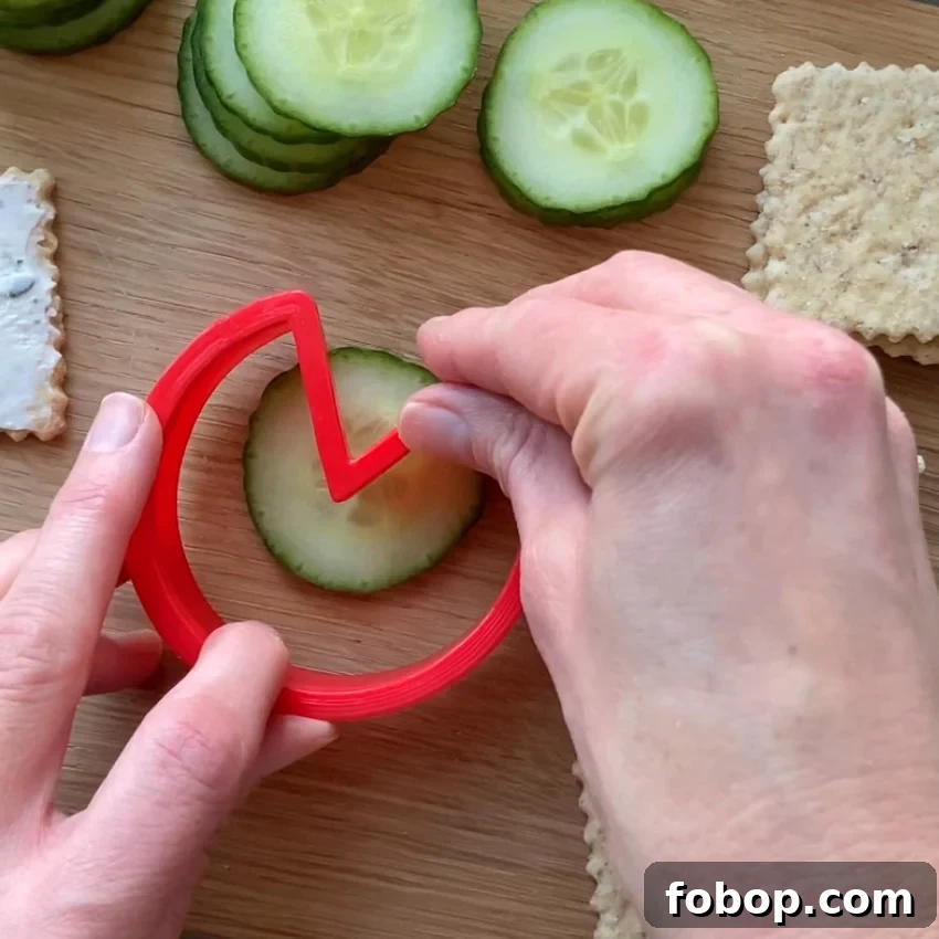A person uses a red zigzag cutter to shape a cucumber slice on a wooden board.