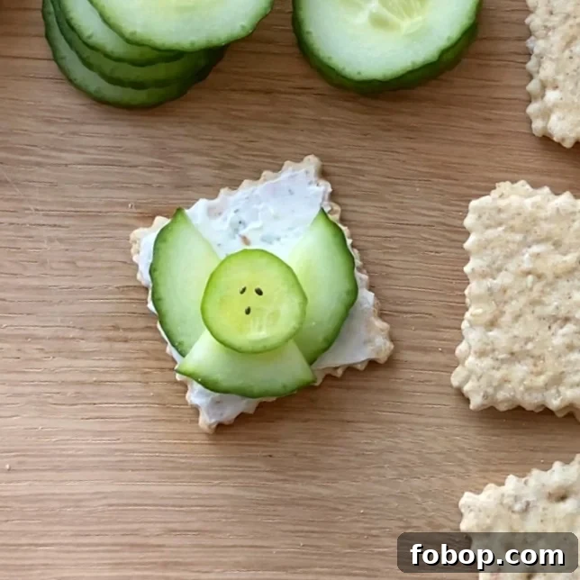 A square cracker with a layer of cream cheese, topped with cucumber slices arranged to resemble an angel.