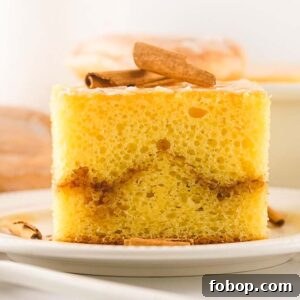 A slice of yellow honey bun cake with a cinnamon swirl, topped with cinnamon sticks, sits on a white plate.
