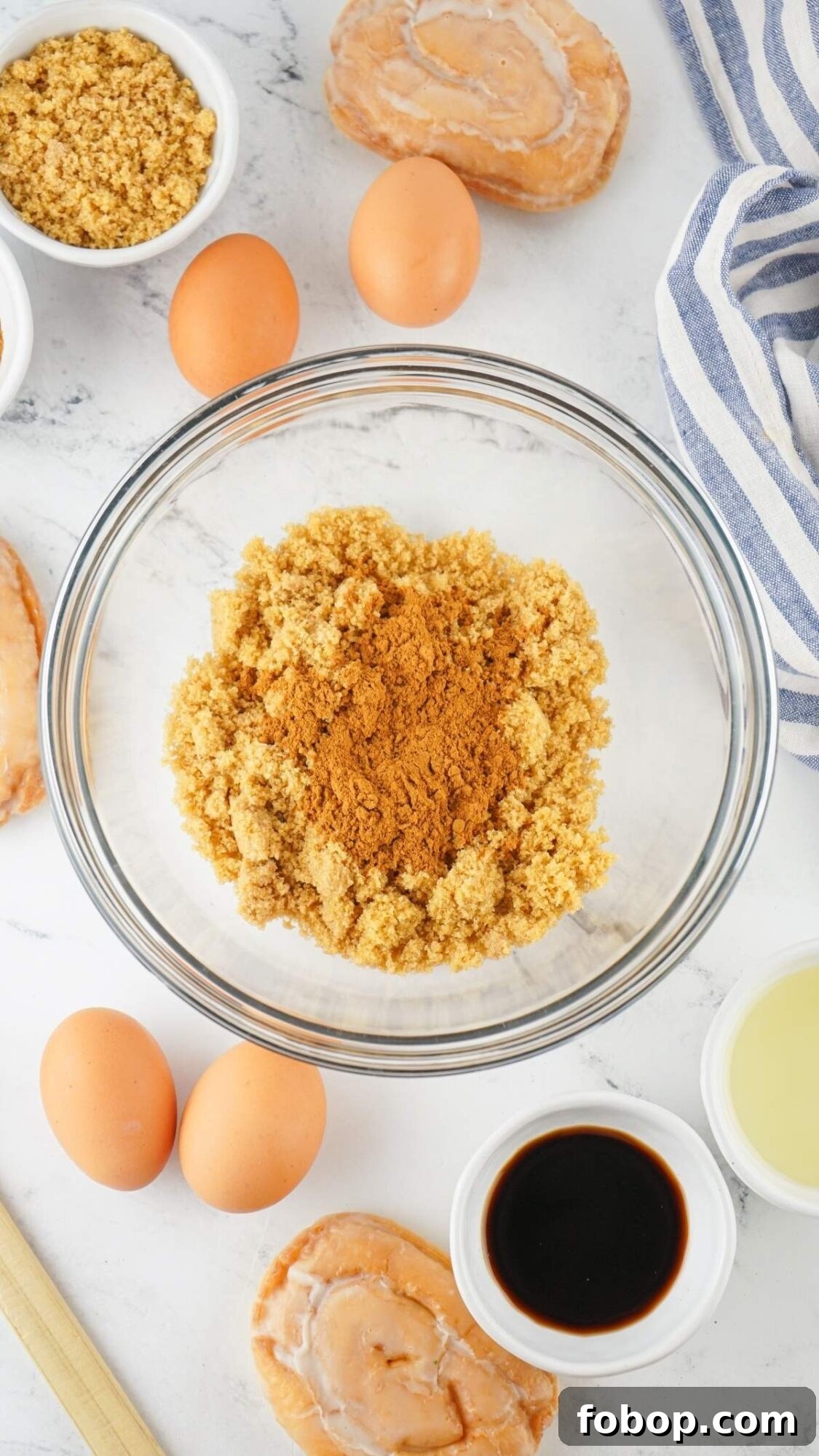 Glass bowl with brown sugar and cinnamon surrounded by ingredients essential for a honey bun cake recipe: eggs, sliced bread, small bowls of vanilla extract and cooking oil, all beautifully arranged on a striped cloth over a white surface.