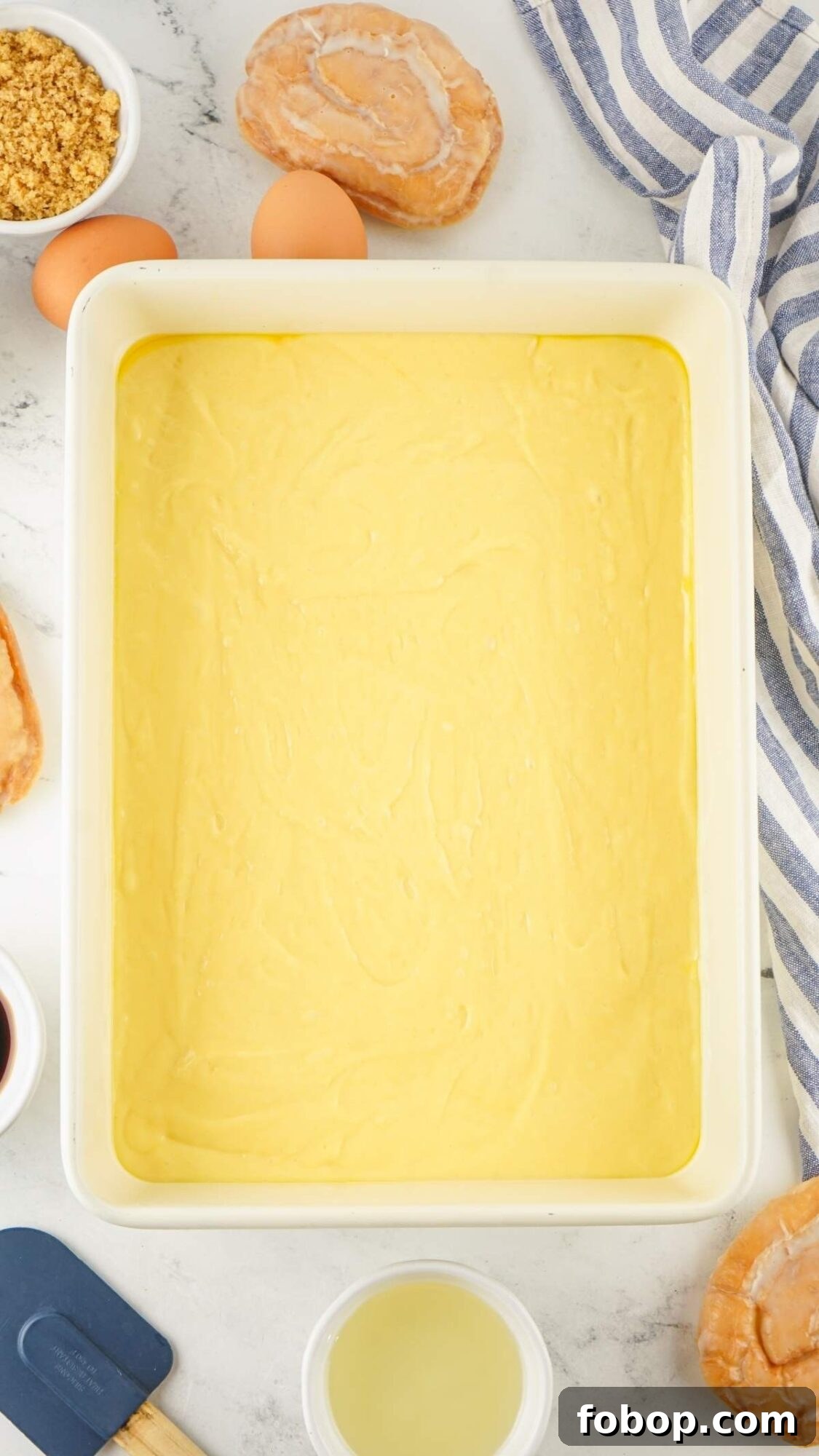 Batter spread evenly in a rectangular baking dish, accompanied by ingredients like eggs, brown sugar, and a striped cloth, creates the perfect honey bun cake recipe.
