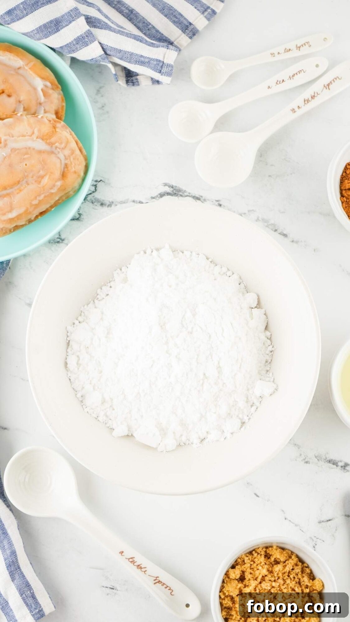 Baking ingredients, including flour, brown sugar, and cinnamon rolls, are arranged on a marble surface. Striped cloths and measuring spoons complete this inviting scene, perfect for a honey bun cake recipe.