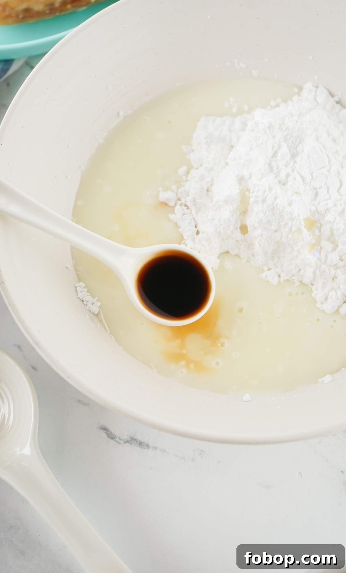 A mixing bowl containing milk, powdered sugar, and a spoon with vanilla extract is ready to create the sweet magic of a honey bun cake.
