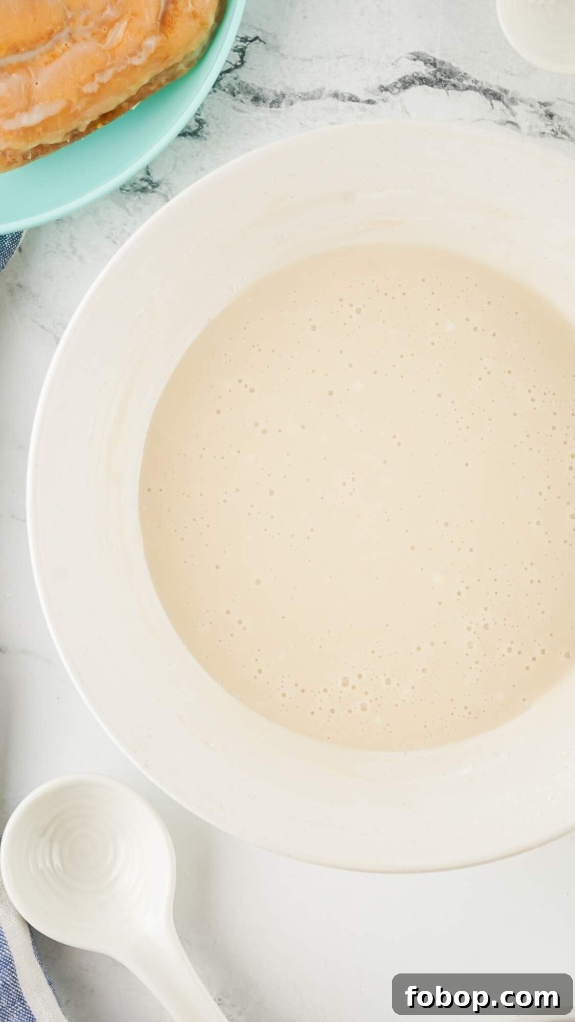 A white bowl filled with a creamy beige liquid, perhaps the start of a honey bun cake recipe, sits on a marble countertop next to a white spoon and a partial view of an orange food item on a turquoise plate.