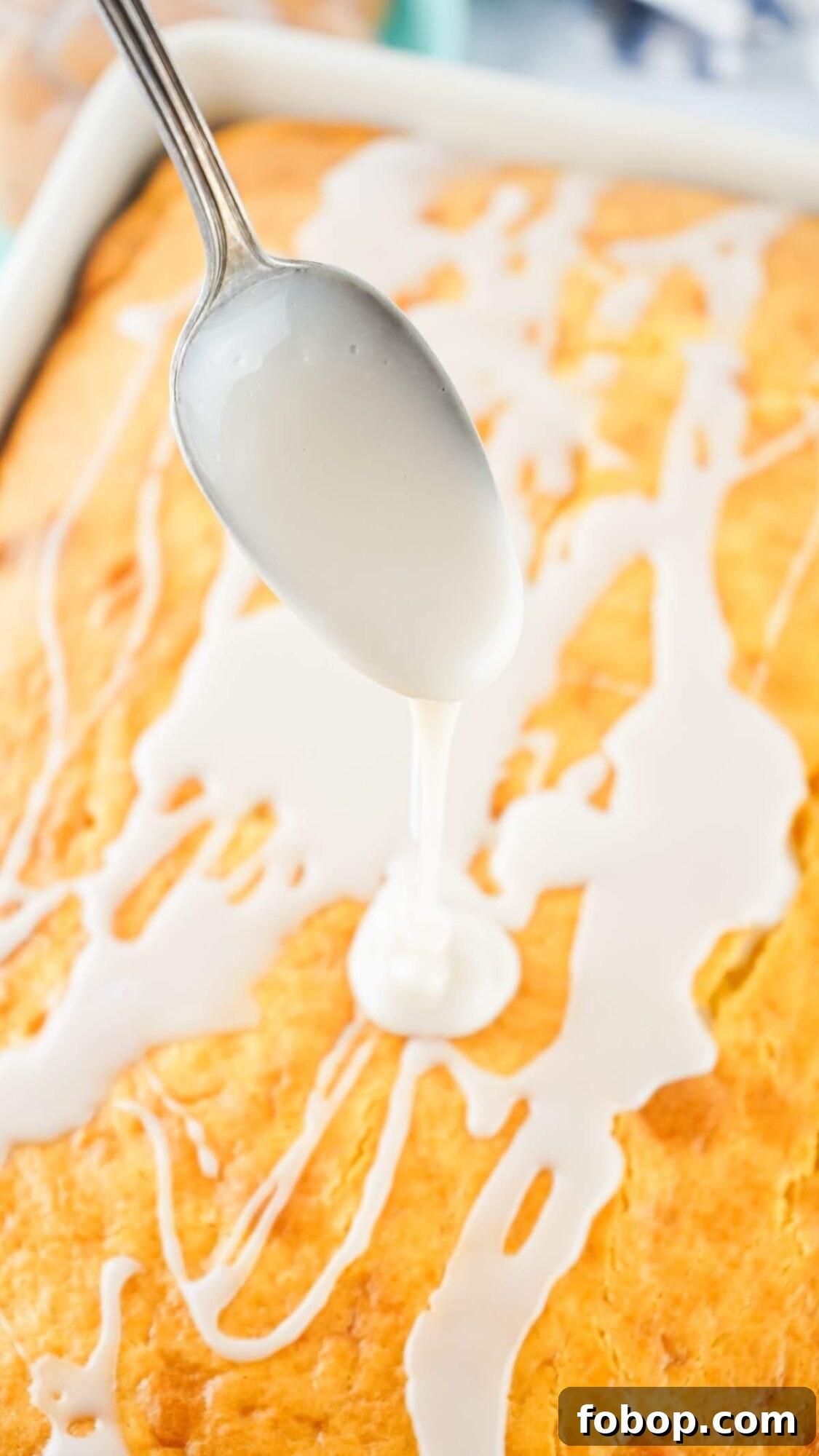 Close-up of a spoon drizzling white icing over a golden-brown honey bun cake, creating a zigzag pattern on the surface.