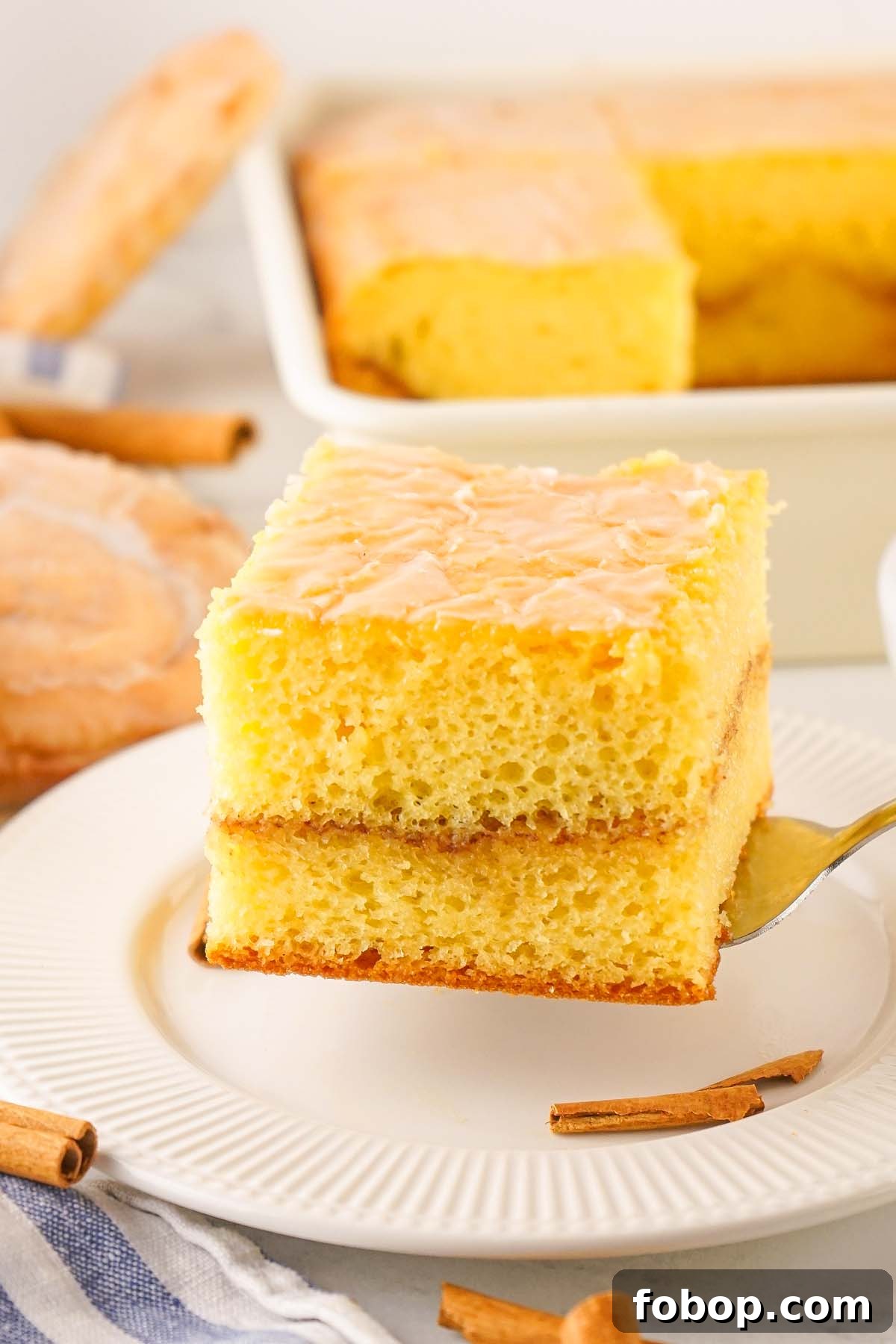 A square slice of yellow honey bun cake recipe with icing is served on a white plate with a fork. A cinnamon stick and a striped napkin are nearby. 