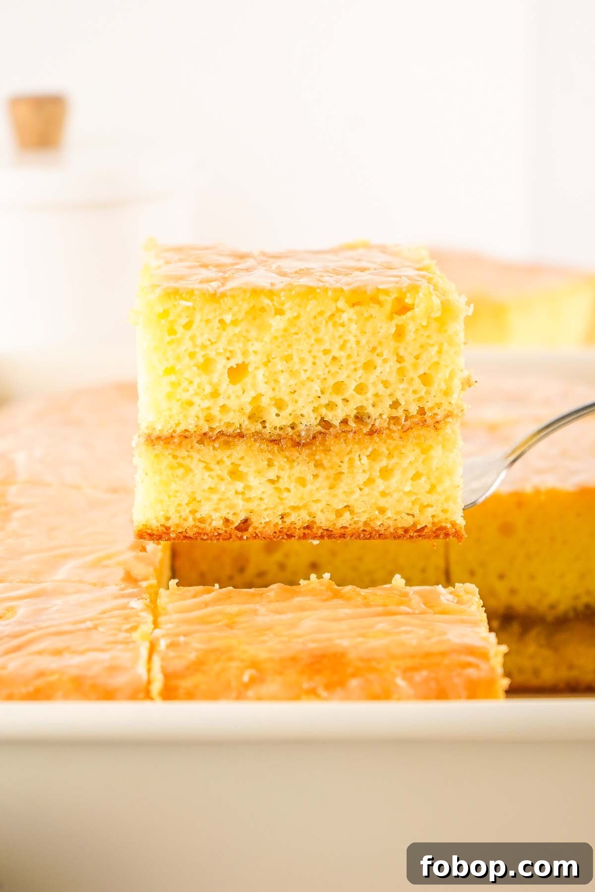 A close-up of a metal spatula lifting a square piece of moist, light-yellow honey bun cake with a shiny glaze from a tray of identical pieces.