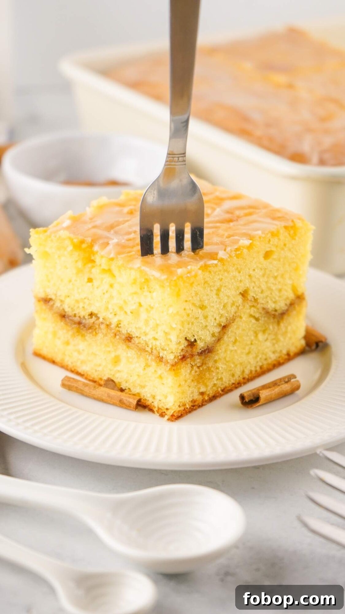 A slice of layered yellow cake (the best honey bun cake) with drizzle and cinnamon sticks, sits invitingly on a white plate accompanied by a fork.