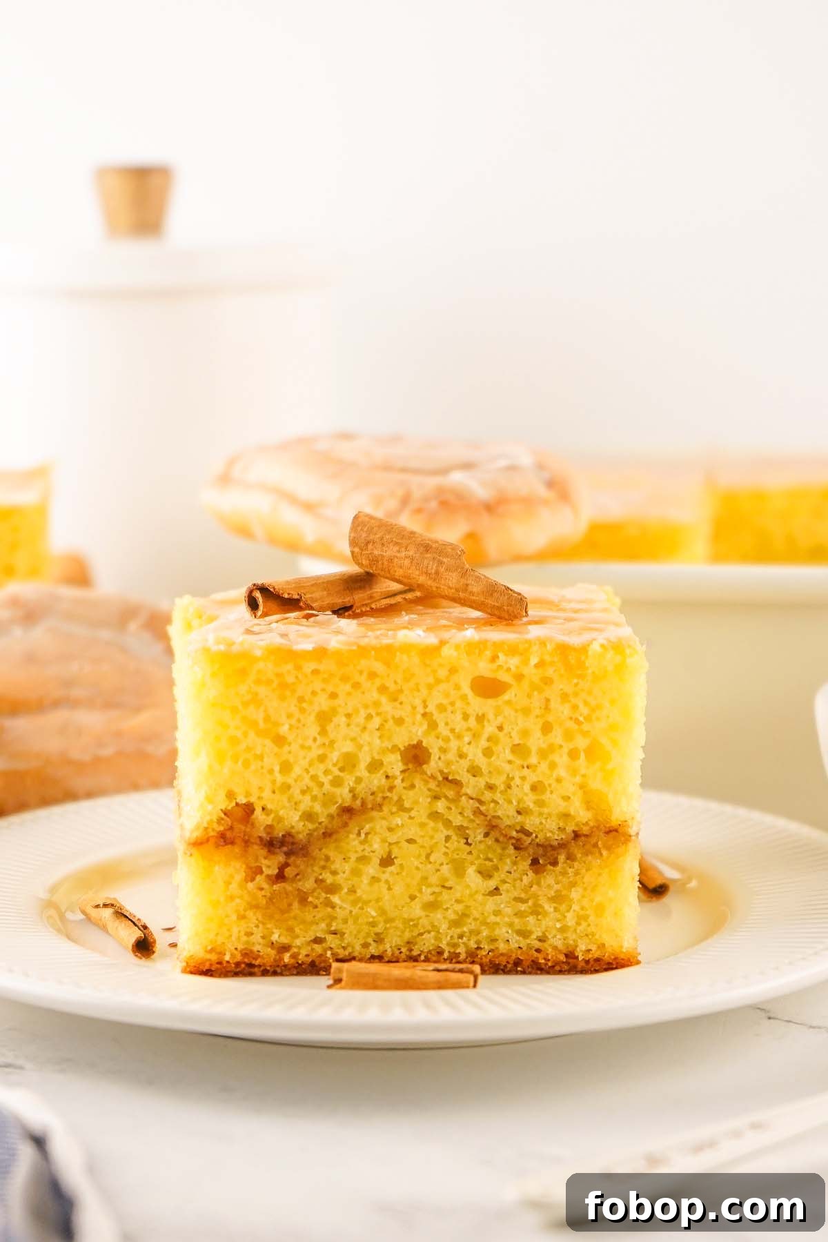 A slice of cinnamon-flavored cake mix honey bun cake, layered with frosting and garnished with cinnamon sticks, is displayed on a white plate.