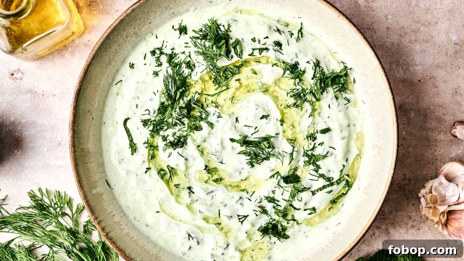 A bowl of creamy tzatziki sauce garnished with chopped dill and a swirl of olive oil. Fresh dill and garlic are nearby.