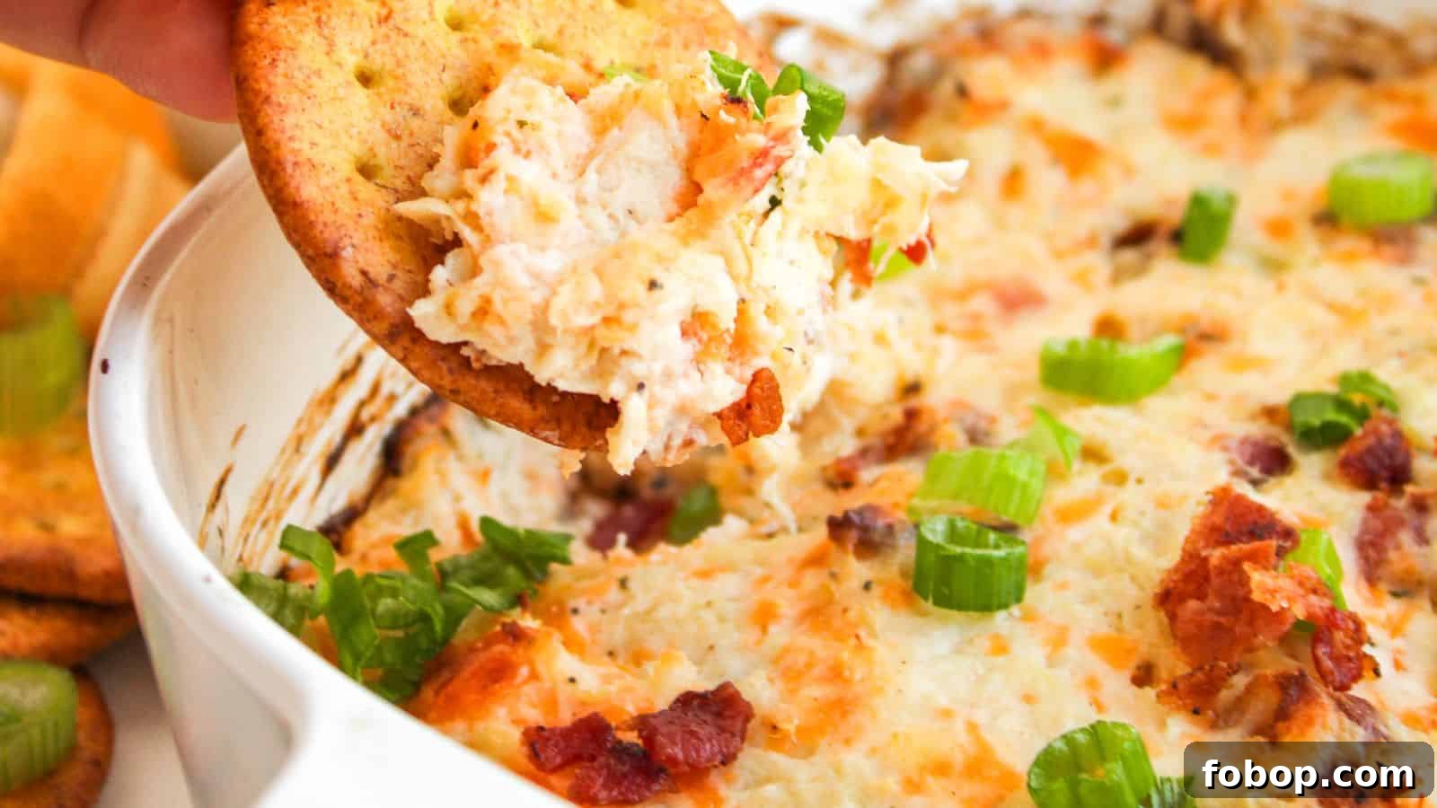 Cracker with creamy dip featuring bacon bits and green onions being lifted from a dish.