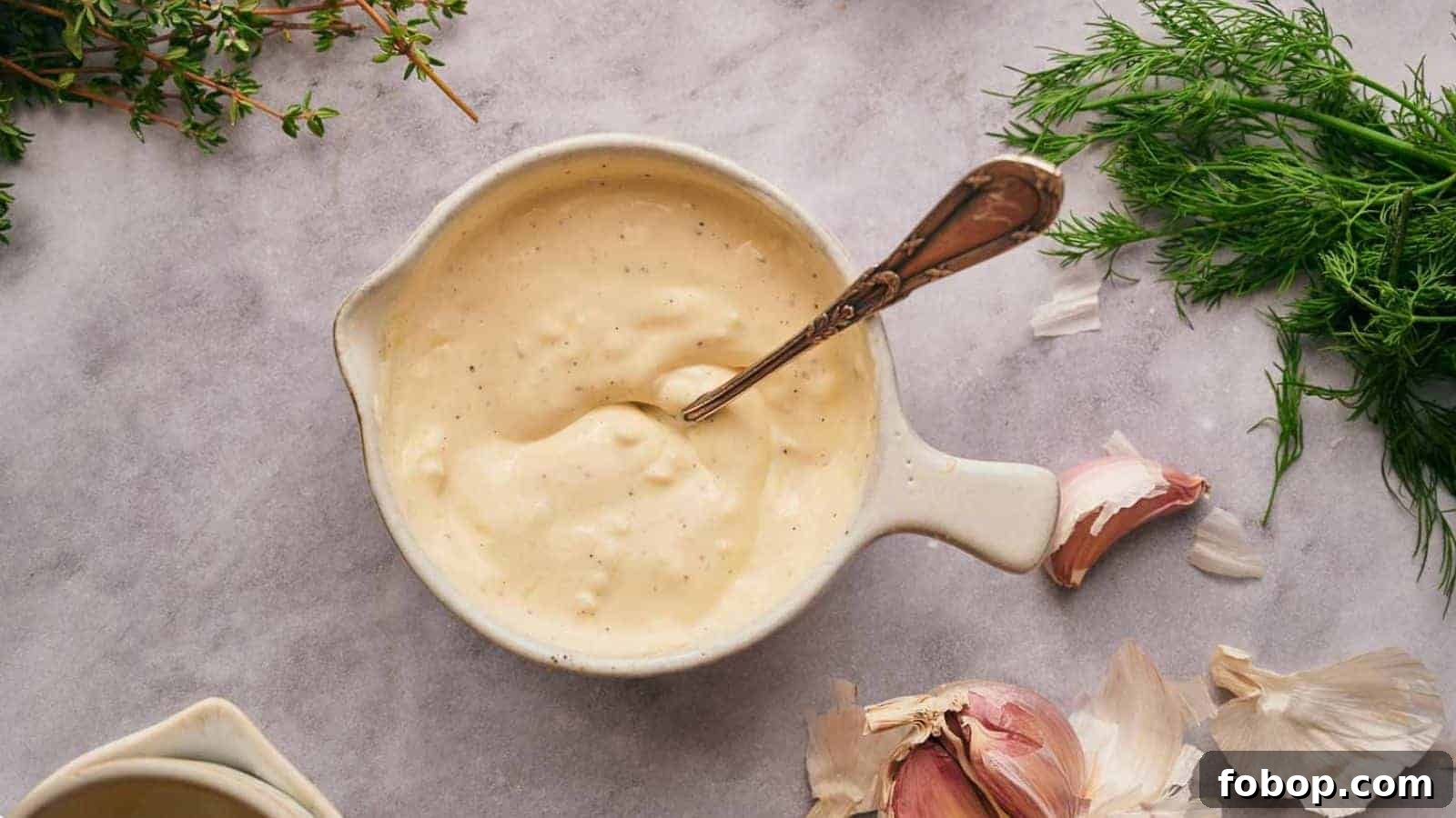 Garlic aioli in a mixing bowl with a spoon in it, garlic cloves, and dill around it.