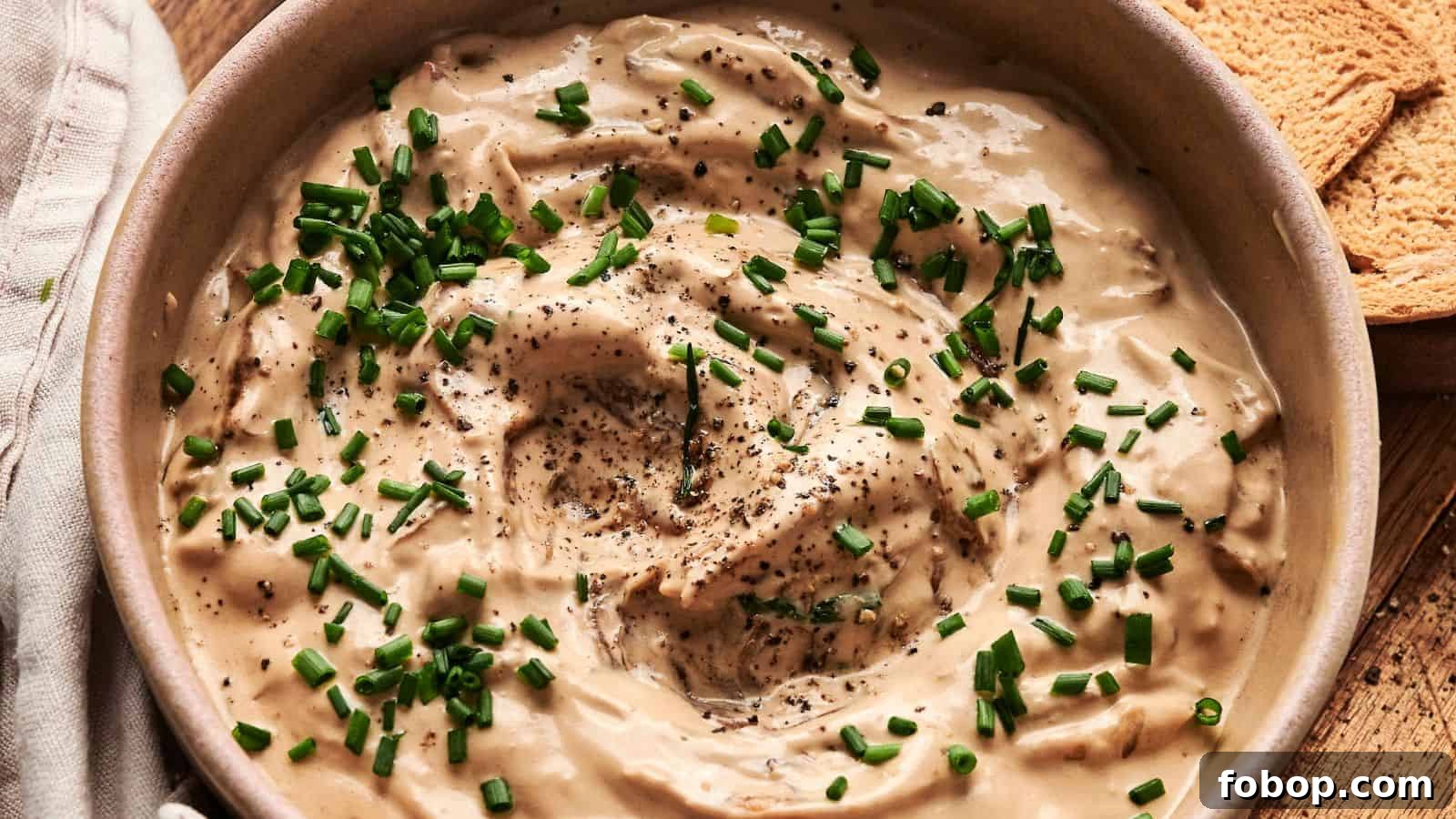 Bowl of creamy dip topped with chopped chives and black pepper, next to pieces of toasted bread.